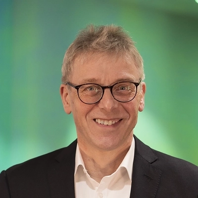 Ein Mann, ca. Mitte 50, mit kurzen grauen Haaren und dunkel gerahmter Brille in weißem Hemd und schwarzem Sakko in einem Büroflur stehend