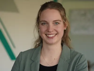 Porträt von Maria Feldhaus, einer jungen Frau mit langem blondem Haar. Sie trägt ein Jackett und lächelt in die Kamera.