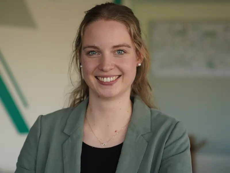 Porträt von Maria Feldhaus, einer jungen Frau mit langem blondem Haar. Sie trägt ein Jackett und lächelt in die Kamera.