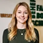 Porträt von Eva Ihlenfeld. Sie trägt langes blondes Haar, ein dunkles Oberteil und lächelt in die Kamera.