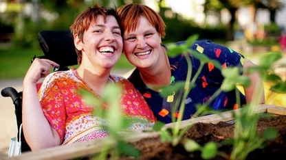 Zwei Frauen in bunt gemusterten Oberteilen sind hinter einem Hochbeet zu sehen. Eine der beiden Frauen sitzt in einem Rollstuhl. Sie stecken die Köpfe zusammen und lachen in die Kamera.