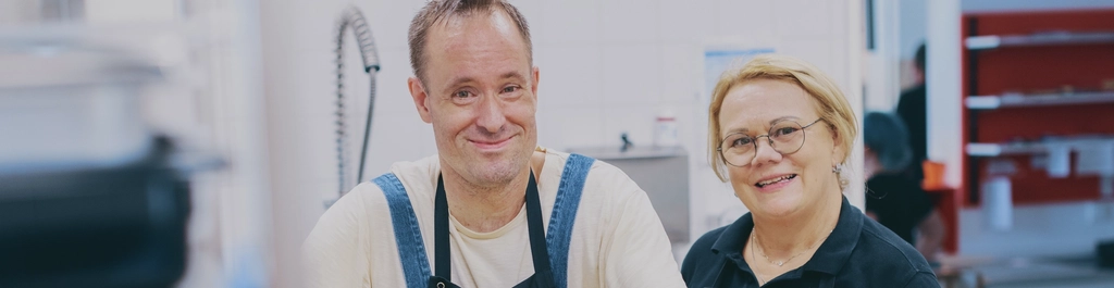 Ein Mann und eine Frau in schwarzer Schürzen stehen in einer Küche. Der Mann trocknet ein gelbes Brett ab.