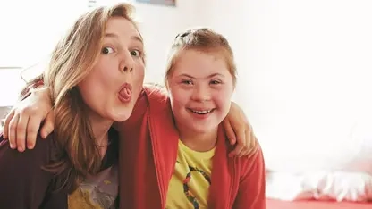 Zwei jugendliche Freundinnen mit und ohne Down-Syndrom Arm in Arm. Eine lacht in die Kamera, die zweite zieht eine lustige Grimasse.