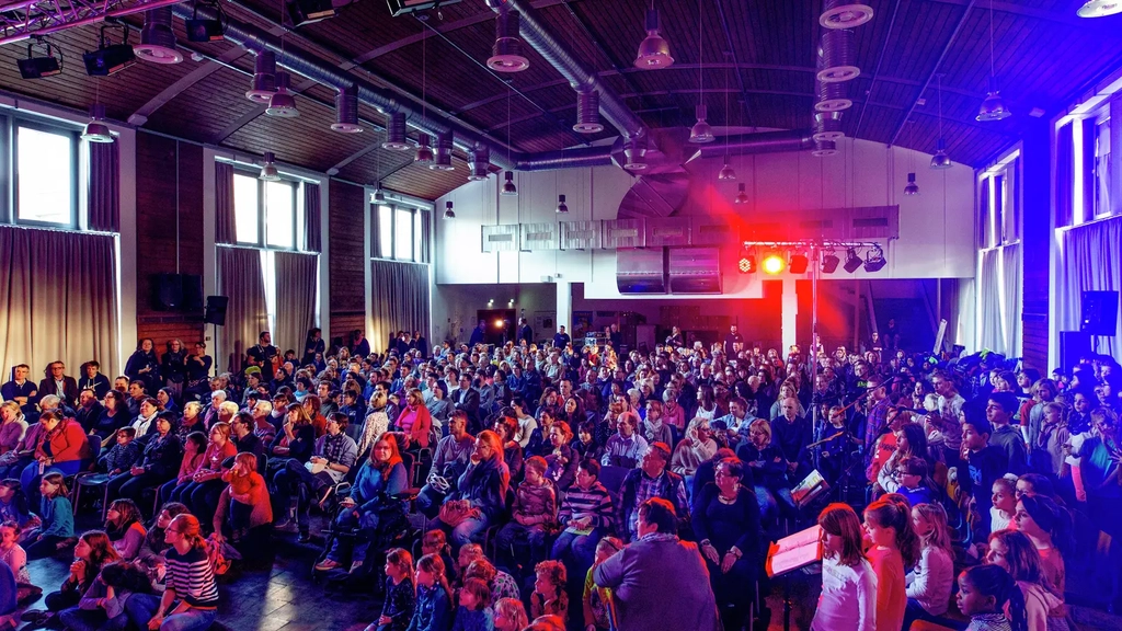 Viele Kinder und Jugendliche sitzen in einer großen, verdunkelten Halle in Reihen und schauen nach vorne. Am Ende des Saals ist ein Scheinwerfer angebracht, der rotes Licht  über die Zuschauer strahlt.