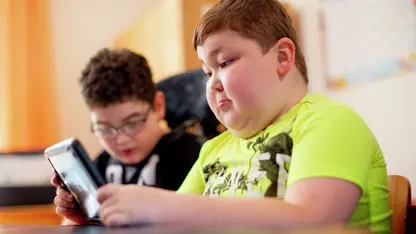 Zwei Jungen sitzen nebeneinander in einem Klassenzimmer. Der Junge im Vordergrund hält ein Tablet in den Händen.