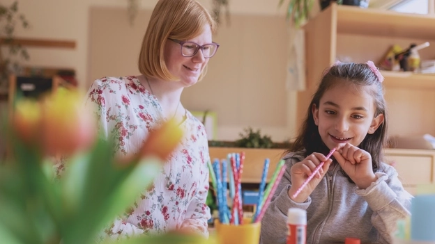 Eine junge Frau mit blonden Haaren, heller geblümter Bluse und Brille sitzt in einem Kita-Raum an einem Basteltisch neben einem dunkelhaarigen ca. fünfjährigem Mädchen mit Migrationshintergrund, die einen Buntstift in der Hand hält.