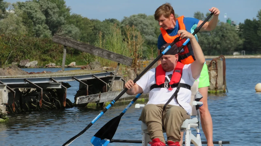 Auf einem Standup-Paddle, auf dem Wsser, sind ein Mann der auf einem Stuhl sitzt, sowie ein stehende Frau zu sehen. Beide halten ein Paddel in der Hand.