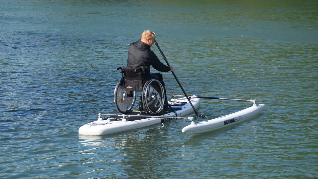 Auf einem besonderen Standup-Paddle, auf dem Wasser, ist ein Mann im Rollstuhl zu sehen, der ein Ruder in der Hand hat.