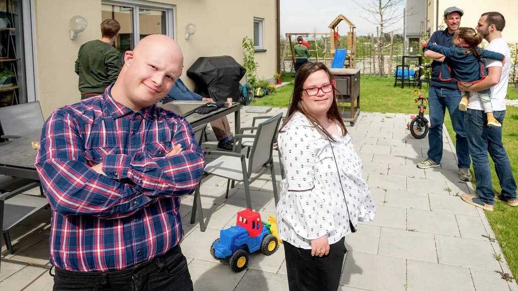 Ein Mann mit cool verschränkten Armen und eine junge Frau mit Brille und Sommerbluse stehen auf der Terrasse ihrer Wohnung. Im Hintergrund zwei junge Väter mit Kind.