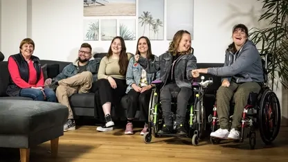 In einem Wohnzimmer sitzen fünf Personen nebeneinander und lachen. Die drei Personen links sitzen auf einem Sofa, die beiden auf der rechten Seite von Rollstühlen, die vor dem Sofa stehen.