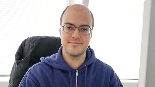 Junger Mann mit dunkler Halbglatze und Brille sitzt in blauer Kapuzenjacke am Schreibtisch vor einem Laptop und großem Monitor..