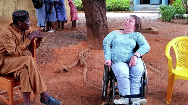 Eine Frau im Rollstuhl und ein schwarzer Mann in traditioneller Kleidung im Gespräch unter einem Baum vor einer afrikanischen Schule.