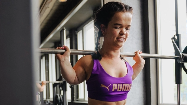 Durchtrainierte kleinwüchsige Frau mit braunen Haaren in Sportkleidung stemmt eine Langhantel auf ihren Schultern