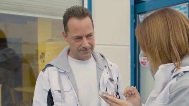 Ein Mann mittleren Alters steht in Malerkleidung neben einer Frau, die ebenfalls Malerkleidung trägt. Sie zeigt ihm etwas auf ihrem Handy, das sie in der Hand hält.