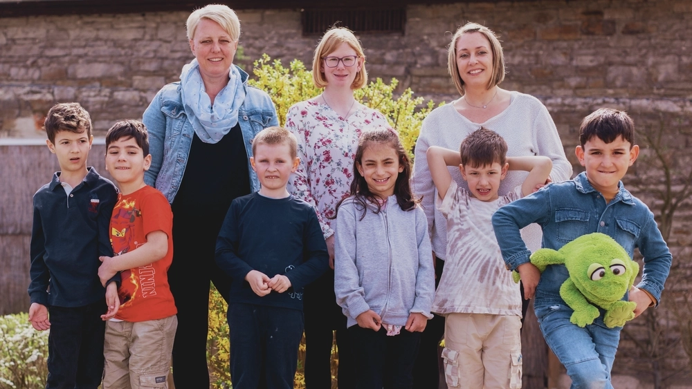 Fünf Jungen und ein Mädchen, alle etwa fünf Jahre alt, stehen in einer Reihe nebeneinander im Garten ihrer Kita. In einer zweiten Reihe dahinter stehen drei Frauen, geschätzt zwischen 30 und 40 Jahren, alle mit blonden Haaren. Die Frau in der Mitte trägt eine helle geblühmte Bluse. Sie ist Kita-Assistentin und hat eine Behinderung, die man ihr aber kaum ansieht.