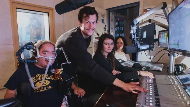Vier Menschen stehen an einem Mischpult. Ein junger Mann sitzt mit einem Beatmungsgerät in einem Rollstuhl.