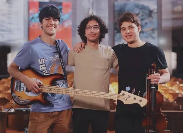 Drei Jugendliche stehen haben jeweils einen Arm und die Schulter des anderen gelegt. Sie halten eine E-Gitarre und eine Geige in der Hand.