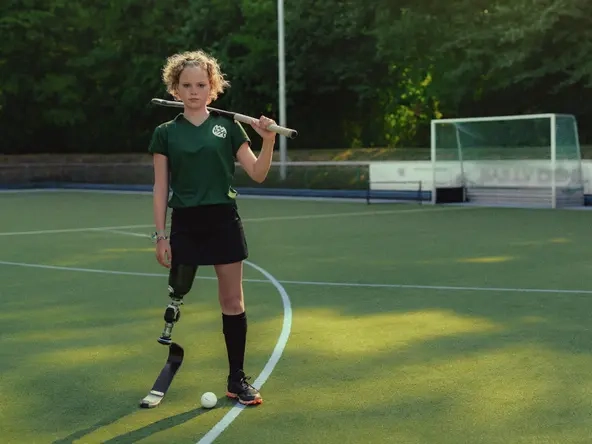 Annika, ein Mädchen mit lockigem Haar und Beinprothese steht auf einem Hockeyplatz. Zwischen ihren Beinen liegt der Ball, ihren Schläger hat sie lässig über die Schultern gelegt.