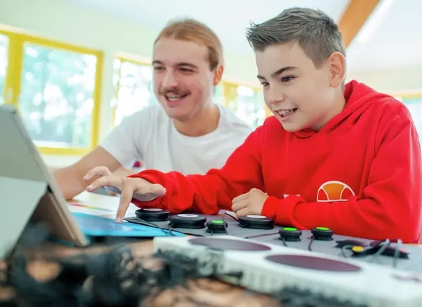 Ein Junge und ein junger Erwachsener sitzen nebeneinander und schauen auf einen Laptop. Vor ihnen liegt ein Controller.
