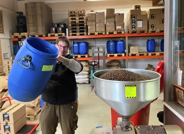 Ein Mann mit Brille schüttet Kaffeebohnen aus einer großen blauen Tonne in einen Trichter