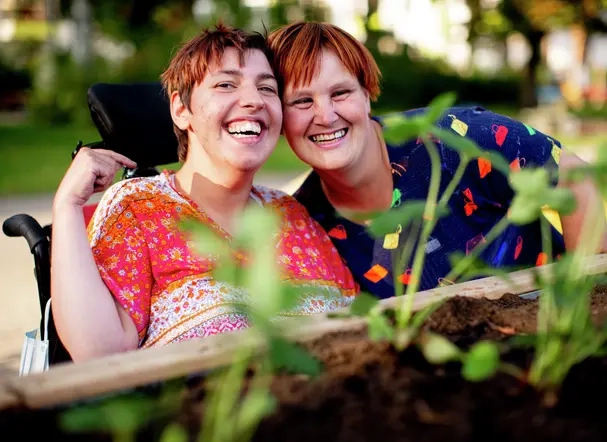 Zwei Frauen in bunt gemusterten Oberteilen sind hinter einem Hochbeet zu sehen. Eine der beiden Frauen sitzt in einem Rollstuhl. Sie stecken die Köpfe zusammen und lachen in die Kamera.