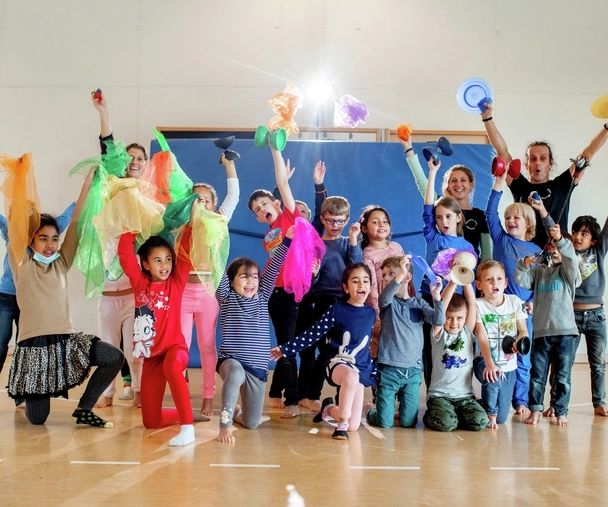 Eine Gruppe von Kindern in einer Turnhalle. Alle jubeln und halten bunte Tücher, Diabolos und Bälle in die Luft.