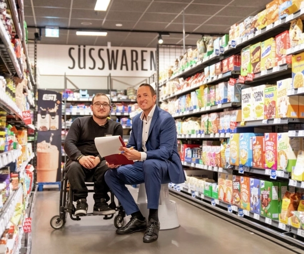 In einem Supermarkt-Gang zwischen zwei Regalreihen sitzen zwei Männer. Links ein Mann in schwarzer Hose und Pulli im Rollstuhl mit Brille und mit Bart, rechts neben ihm ein Mann mit kurzen dunklen Haaren in blauem Anzug und weißem Hemd auf einem Tritthocker. Beide lächeln in die Kamera.