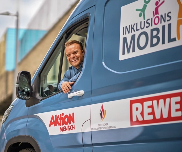 In einem blauen Kleintransporter sitzt ein kleinwüchsiger, sportlicher Mann mit kurzen blonden Haaren. Er sieht aus dem Fenster der Fahrertür und lacht in die Kamera. Auf dem Bus sind Ausfkleber angebracht. Auf einem steht "Inklusionsmobil". Auf den anderen sind die Logos der kooperatiosnpartner zu sehen, die diesen Bus unterstützen: Aktion Mensch, Deutscher Behindertensportverband und REWE.