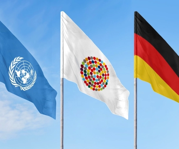 Drei Flaggen an Mast vor blauem Himmel: Eine mit dem Logo der UN, eine mit vielen bunten Punkten im Kreis, die Inklusion symbolisieren sollen und die deutsche Fahne