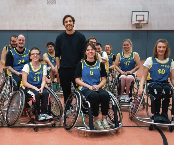 Max Giesinger steht in einer Turnhalle in mitten einer Gruppe Rollstuhl-Basketballer*innen