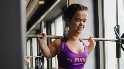 Durchtrainierte kleinwüchsige Frau mit braunen Haaren in Sportkleidung stemmt eine Langhantel auf ihren Schultern