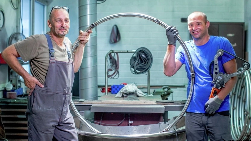 Zwei Männer stehen in einer Werkstatt lächelnd nebeneinander und tragen gemeinsam einen Reifen aus Metall