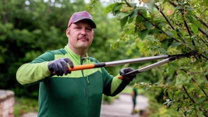 Ein Mann trägt eine rote Kappe, eine grüne Jacke und Gartenhandschuhe. Er hat eine große Baumschere in der Hand und schneidet einen Busch.