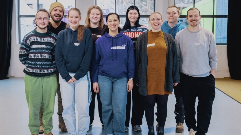 ein Gruppe von 9 Jugendlichen und jungen Erwachsenen steht zum Gruppenbild in einem großen Studio-Raum. Sie sind locker leger gekleidet. Auf der Brust haben si eKlebestreifen, auf denen ihr Name geschrieben steht.