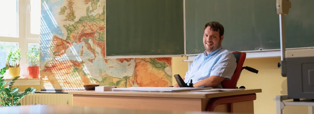 Ein junger Mann in einem Elektrorollstuhl sitzt hinter einem Lehrerpult in einem Klassenzimmer. Hinter ihm seind eine große Landkarte und die Tafel zu sehen.