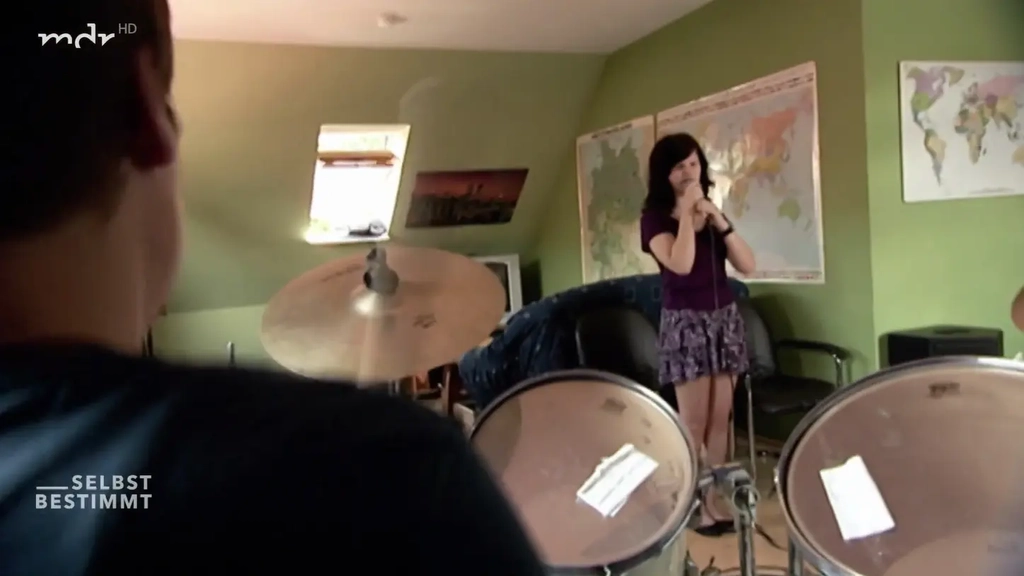 Die 13-jährige Marie Lampe (ein Mädchen mit langen dunklen Haaren in T-shirt und Rock) steht in einem grün gestrichenen Zimmer und singt in ein Mikrofon. An der Wand hängen Weltkarten. Im Vordergrund sieht man unscharf ein Schlagzeug und einen Jungen.