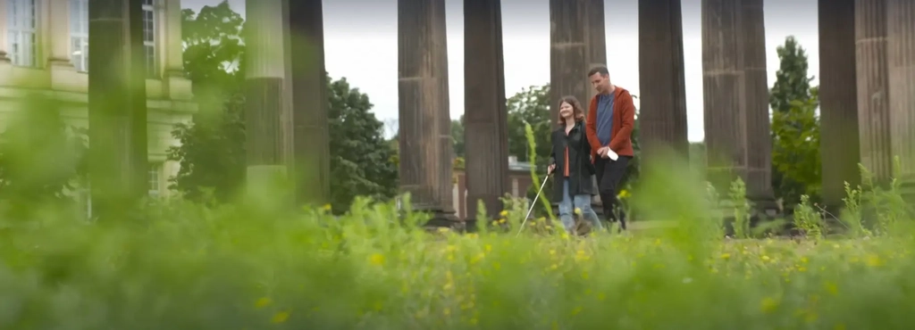Eine junge Frau mit Blindenstock und ein junger Mann gehen spazieren. Im Vordergrund ist Wiese zu sehen. Im Hintergrund Säulen der Universität Potsdam.