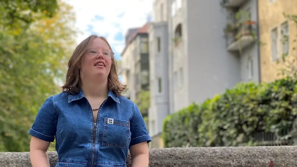 Eine junge Frau mit Down-Syndrom lehnt lässig an einer Mauer. Sie hat schulterlange blonde Haare, trägt eine Brille und einen blauen Hosenanzug aus Jeansstoff, und Sneaker mit Leopardenfell-Muster. Im Hintergrund Hausfassaden und Bäume.