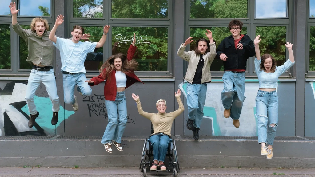 Eine diverse Gruppe von Jugendlichen vor einer Schulfassade, unter dessen Fenster Graffiti an der Wand ist. In der Mitte der Gruppe eine Rollifahrerin mit nach oben gereckten Armen. Links und rechts von ihr springen je drei junge Menschen von einem kleinen Absatz auf die Erde.
