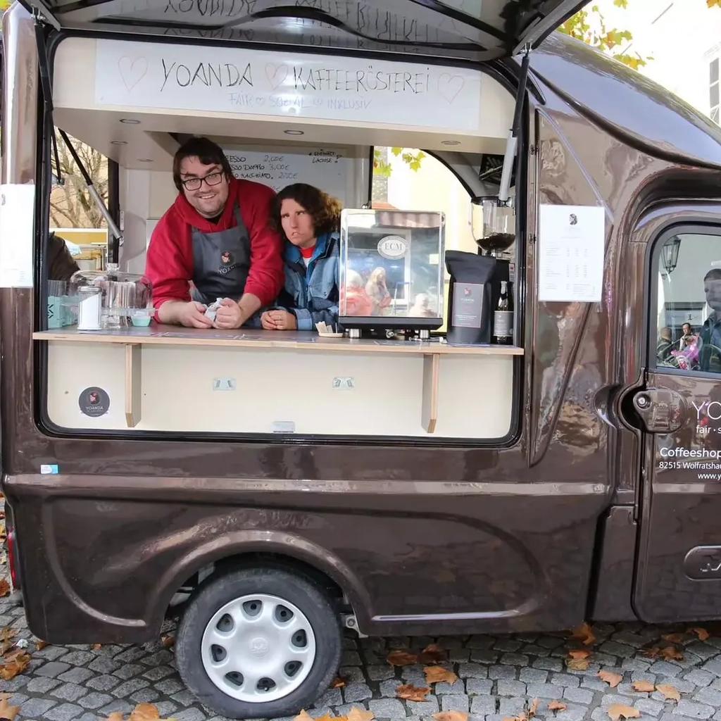 Ein Mann und eine Frau mit Schürze stehen in einem Zum Kaffee-Roller umgebauten dunklen Kastenwagen in einer Fußgängerzone in der Münchner Innenstadt.