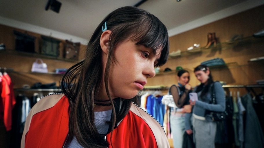 Eine junge Frau mit Downsyndrom steht in einem Bekleidungsgeschäft. Sie schaut unsicher über ihre linke Schulter. Im Hintergrund sind zwei weitere junge Frauen erkennbar, die scheinbar miteinander tuscheln.