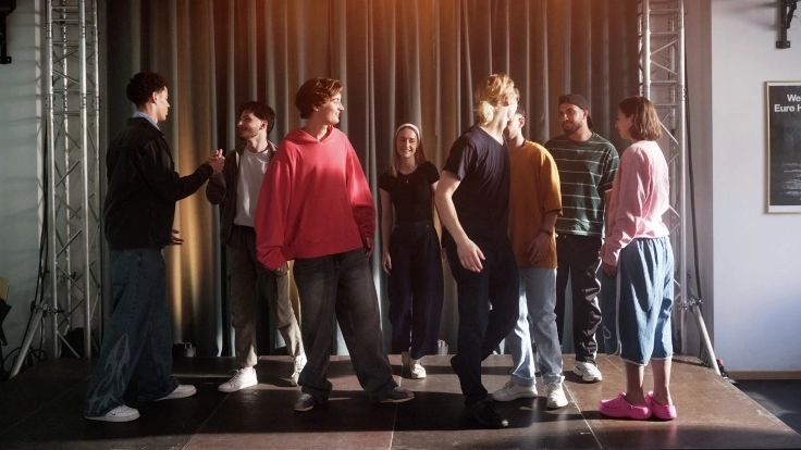 Eine Gruppe Jugendlicher steht auf einer Bühne. Sie schlagen miteinander ein und schauen sich freundlich gegenseitig an.