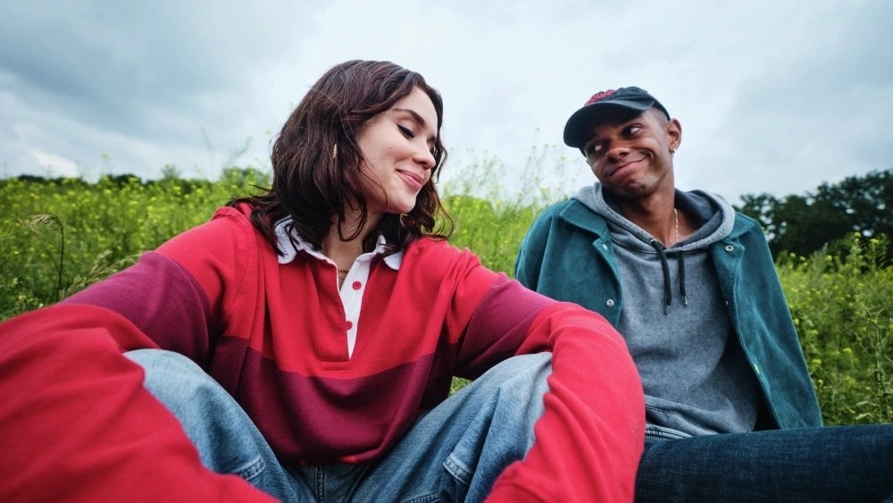 Zwei Jugendliche sitzen draußen auf einer Wiese. Das Mädchen auf der linken Seite trägt ein rotes Langarmshirt und Jeans, der Junge auf der rechten Seite eine graue Kapuzenjacke unter einer blauen Jacke sowie eine Baseballkappe. Sie schauen einander freundschaftlich an.