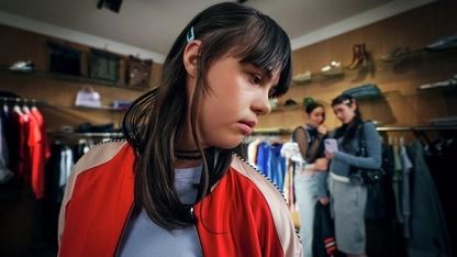 Eine junge Frau mit Downsyndrom steht in einem Bekleidungsgeschäft. Sie schaut unsicher über ihre linke Schulter. Im Hintergrund sind zwei weitere junge Frauen erkennbar, die scheinbar miteinander tuscheln.