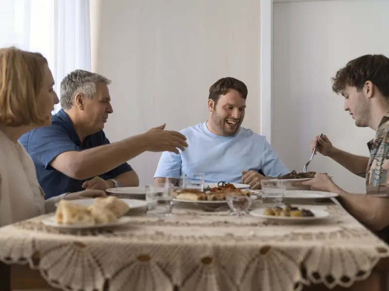 Eine Familie mit vier Personen sitzt an einem Tisch und isst gemeinsam. Sie besteht aus zwei jungen Männern sowie Mutter und Vater. Einer der beiden jungen Männer sitzt in einem Elektrorollstuhl.