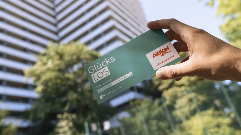Eine Hand hält ein Glücks-Los der Aktion Mensch in die Luft. Im Hintergrund sieht man ein  Hochhaus.