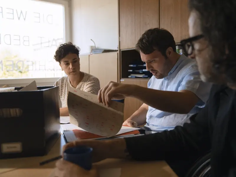 Drei Personen sitzen gemeinsam in einem Lehrerzimmer und schauen sich Akten an. Einer der drei sitzt in einem Elektrorollstuhl.