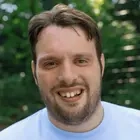 Ein junger mann mit mittelbraunem Haar und hellem T-Shirt schaut in die Kamera und lächelt.