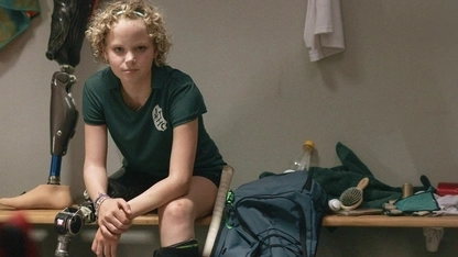 Ein blondes Mädchen sitzt in einer Umkleidekabine. Sie trägt ein grünes Trikot, neben ihr steht an Hockeyschläger. Sie hat eine Prothese am rechten Bein und schaut entschlossen in die Kamera.