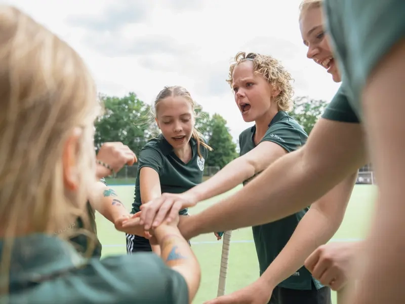 Eine Gruppe von Mädchen in grünen Trikots steht auf einem Hockeyplatz. Sie haben sich im Kreis aufgestellt und ihre Hände in der Mitte übereinandergelegt. Ein Mädchen mit blonden Locken hat den Mund leicht geöffnet und schaut ernst, als ob sie mit den anderen spricht.